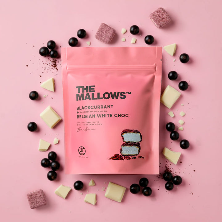 Pink package of 'The Mallows' blackcurrant Belgian white choc on a pink background with blackcurrants and white chocolate pieces.