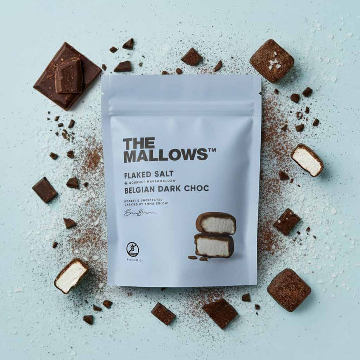 Packaging of 'The Mallows' flaked salt Belgian dark choc marshmallows on a light blue background with scattered chocolate pieces.